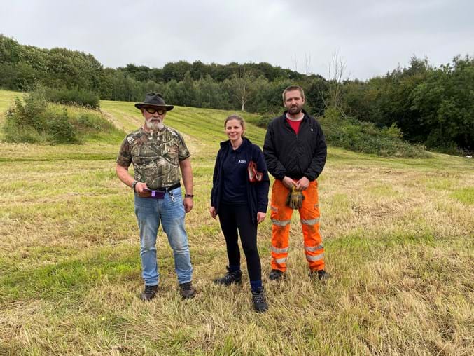 Three volunteers stood in a cut wildflower meadow, green landscape with trees in the background.